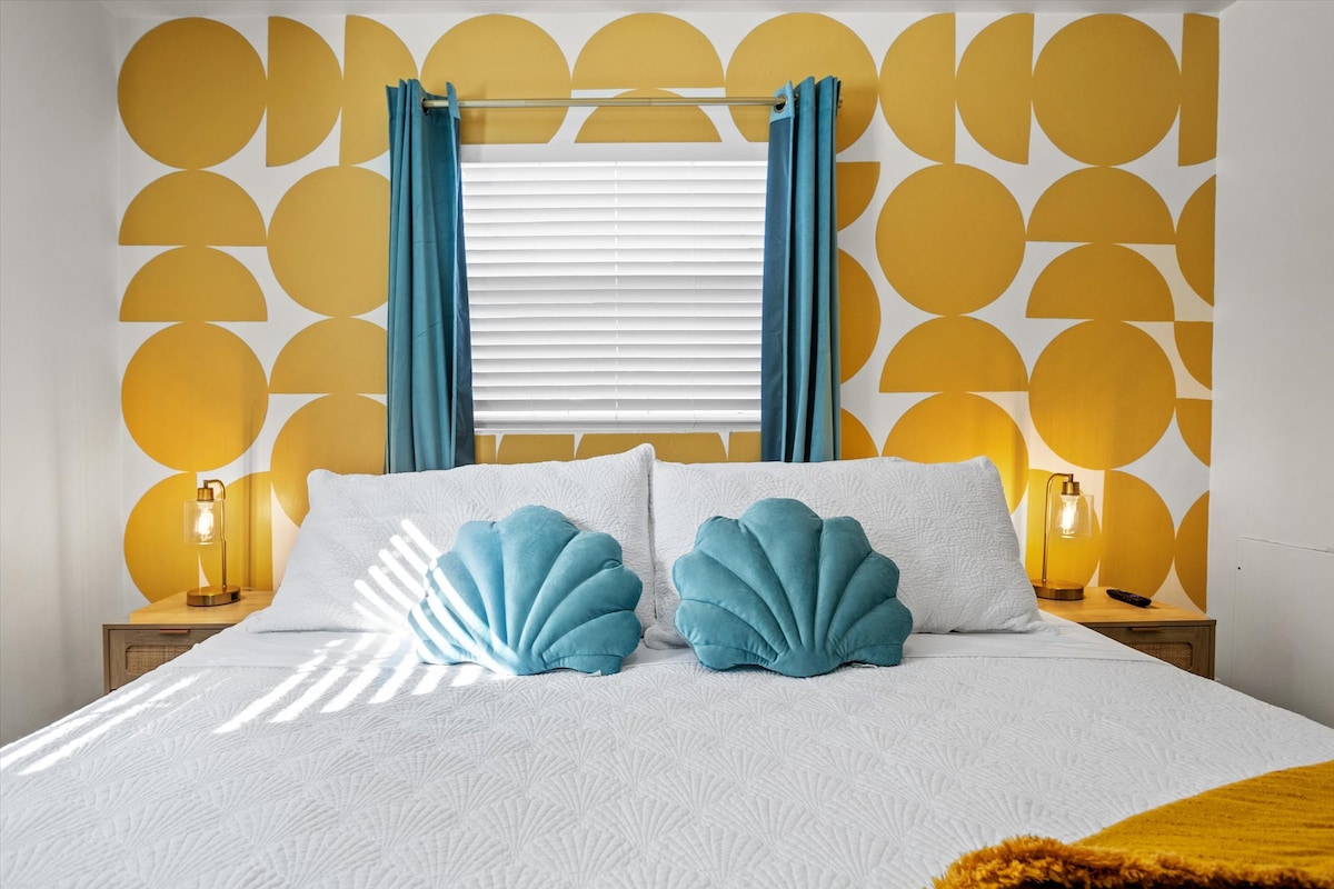 A cozy bedroom features a spacious bed with a white quilted coverlet and decorative turquoise shell-shaped pillows. The wall behind showcases a vibrant yellow circular pattern, complemented by golden bedside lamps on wooden nightstands. Natural light filters through a window adorned with light blue curtains.