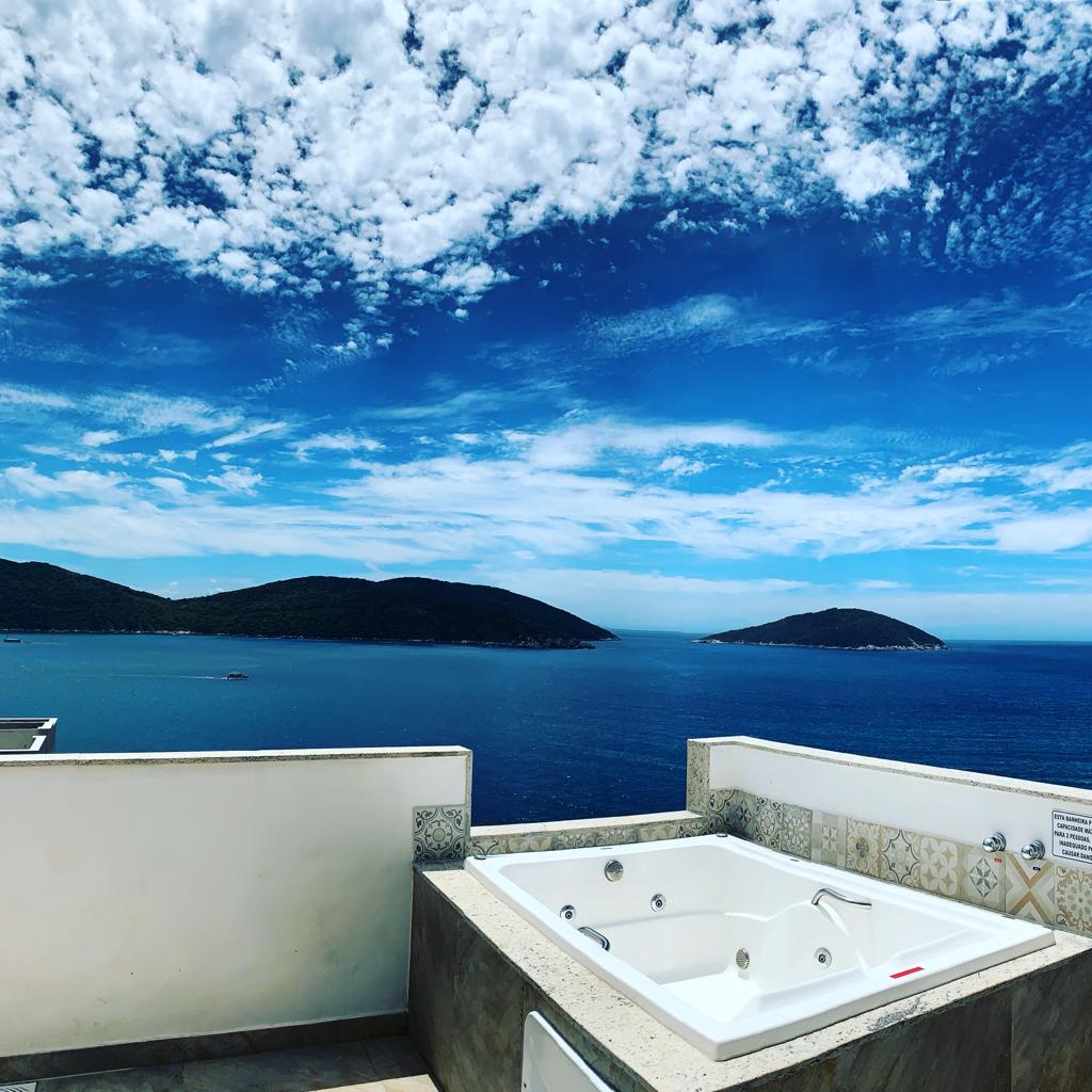 An outdoor hot tub is positioned on a patio overlooking expansive ocean views. The clear blue sky is adorned with clouds, while distant islands are visible on the horizon. The tub's neutral stone surround complements the tranquil waterfront setting.