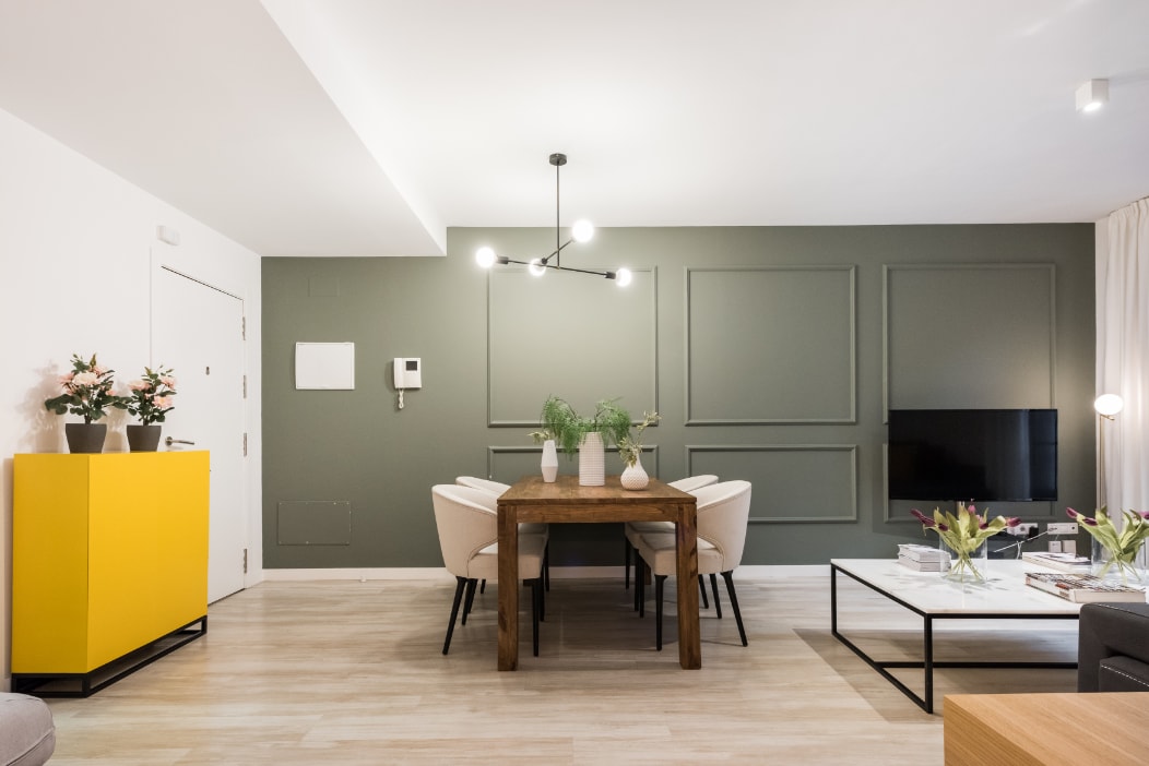 A dining area is depicted with a wooden table surrounded by four light-colored chairs. A striking yellow cabinet stands against the wall, complemented by two potted plants atop the table. A modern light fixture hangs above, and a wall-mounted television is visible.