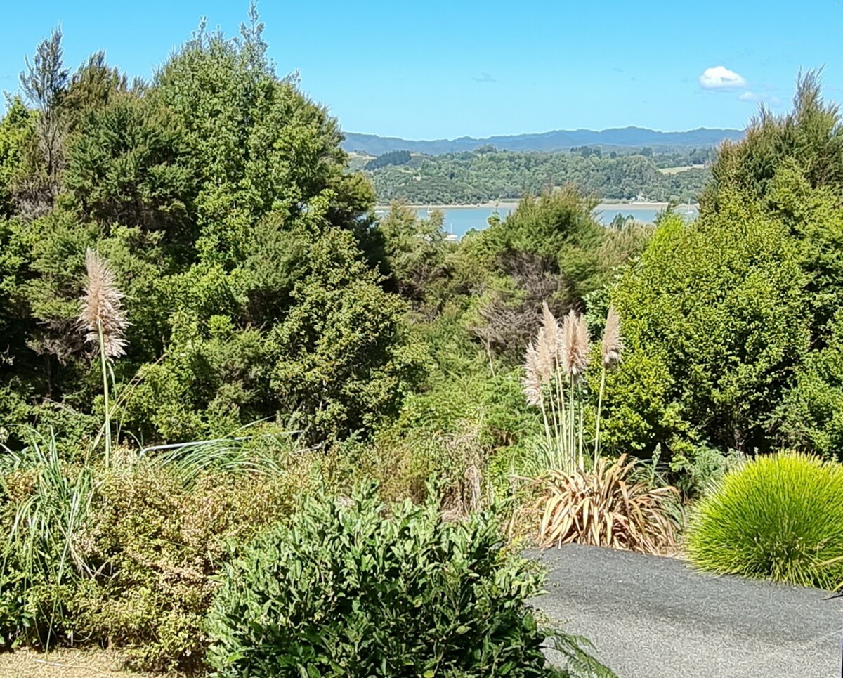 Lush greenery and native foliage create a serene foreground, while a tranquil body of water and distant hills are visible in the background. The scene is framed by a clear blue sky, enhancing the natural beauty of the surroundings.
