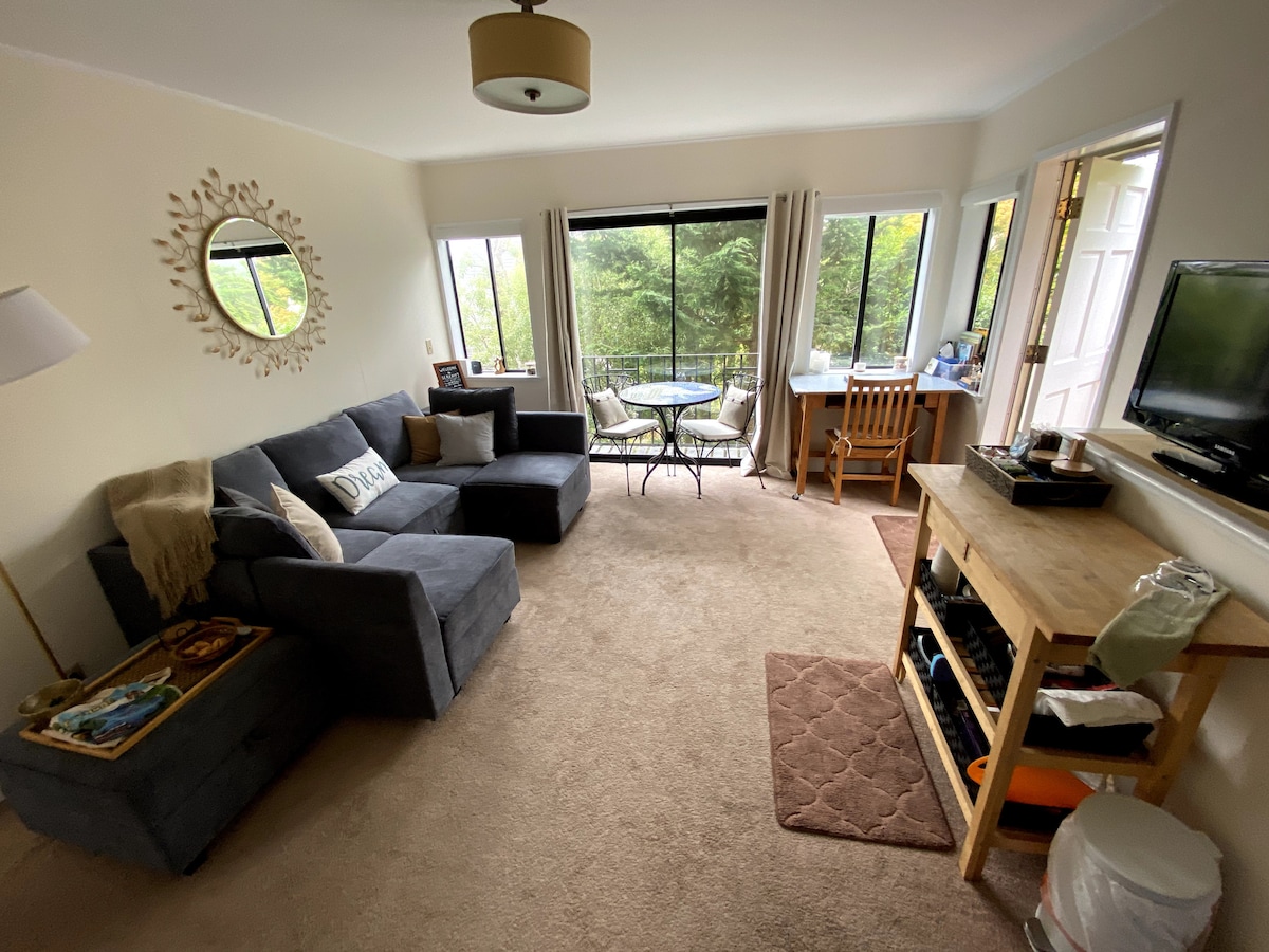 The cozy living room features a sectional sofa, a wall-mounted flat screen TV, and a wooden console table. A cafe table with two chairs is positioned near large windows that offer views of the outdoors, bringing natural light into the space.