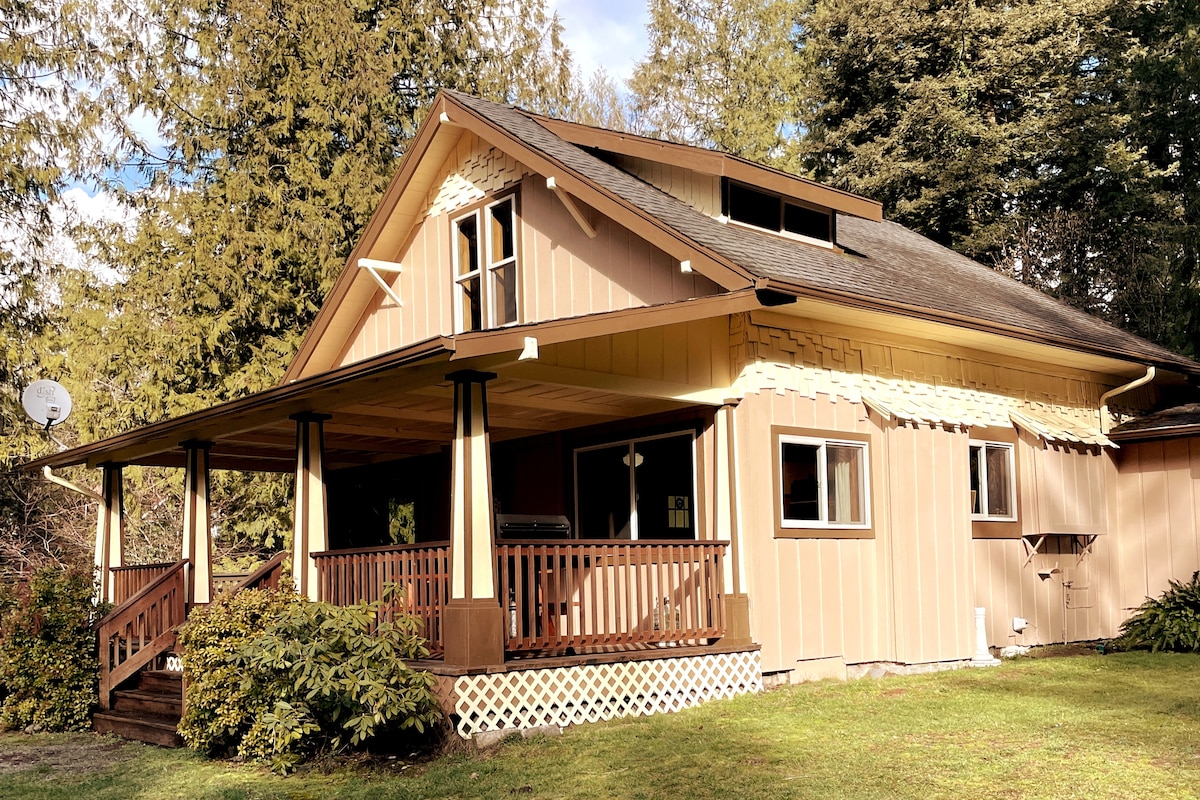 The exterior of a spacious lodge is shown, featuring a welcoming porch with wooden railings and columns. The house is set against a backdrop of trees, with large windows allowing for natural light. The roof is pitched, and a small satellite dish is mounted on the side.