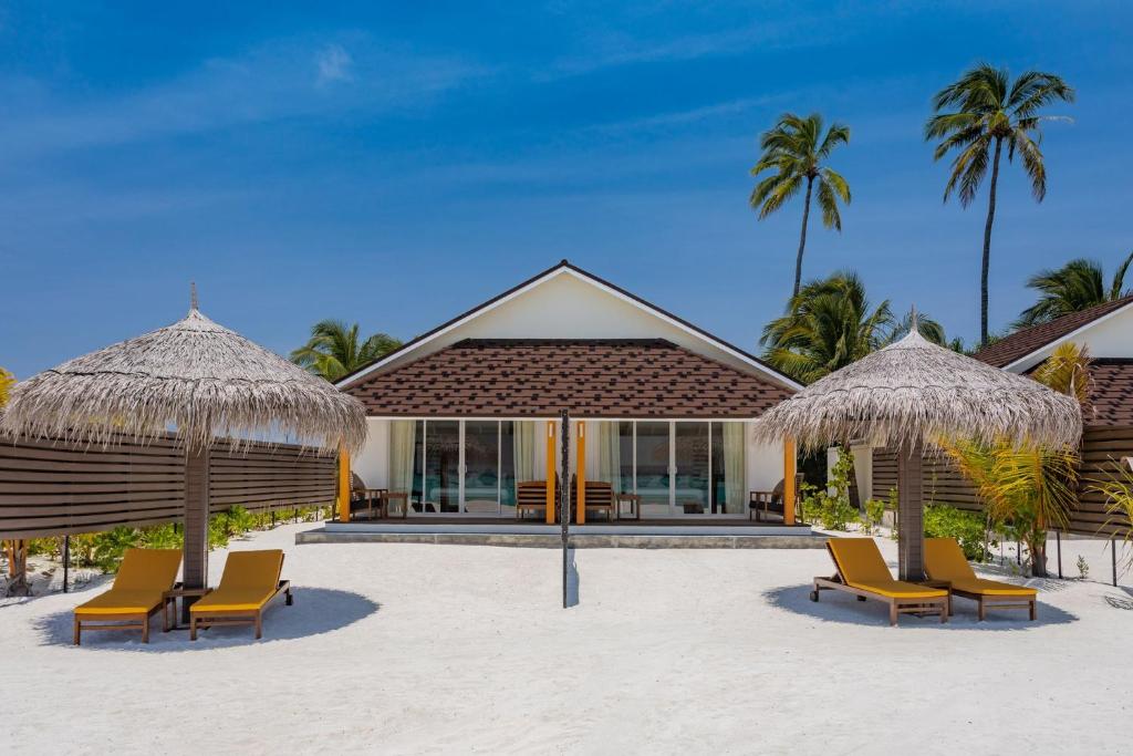 A beachfront villa is framed by two shaded cabanas, each with comfortable lounge chairs. The structure features large glass doors that provide direct access to the sandy beach, with palm trees gently swaying in the background under a clear blue sky.