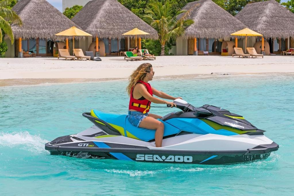 A jet ski is seen gliding over the clear turquoise water near a sandy beach. In the background, thatched-roof cabanas with yellow umbrellas provide shaded seating, inviting relaxation. Lush greenery frames the scene, creating a vibrant coastal atmosphere.
