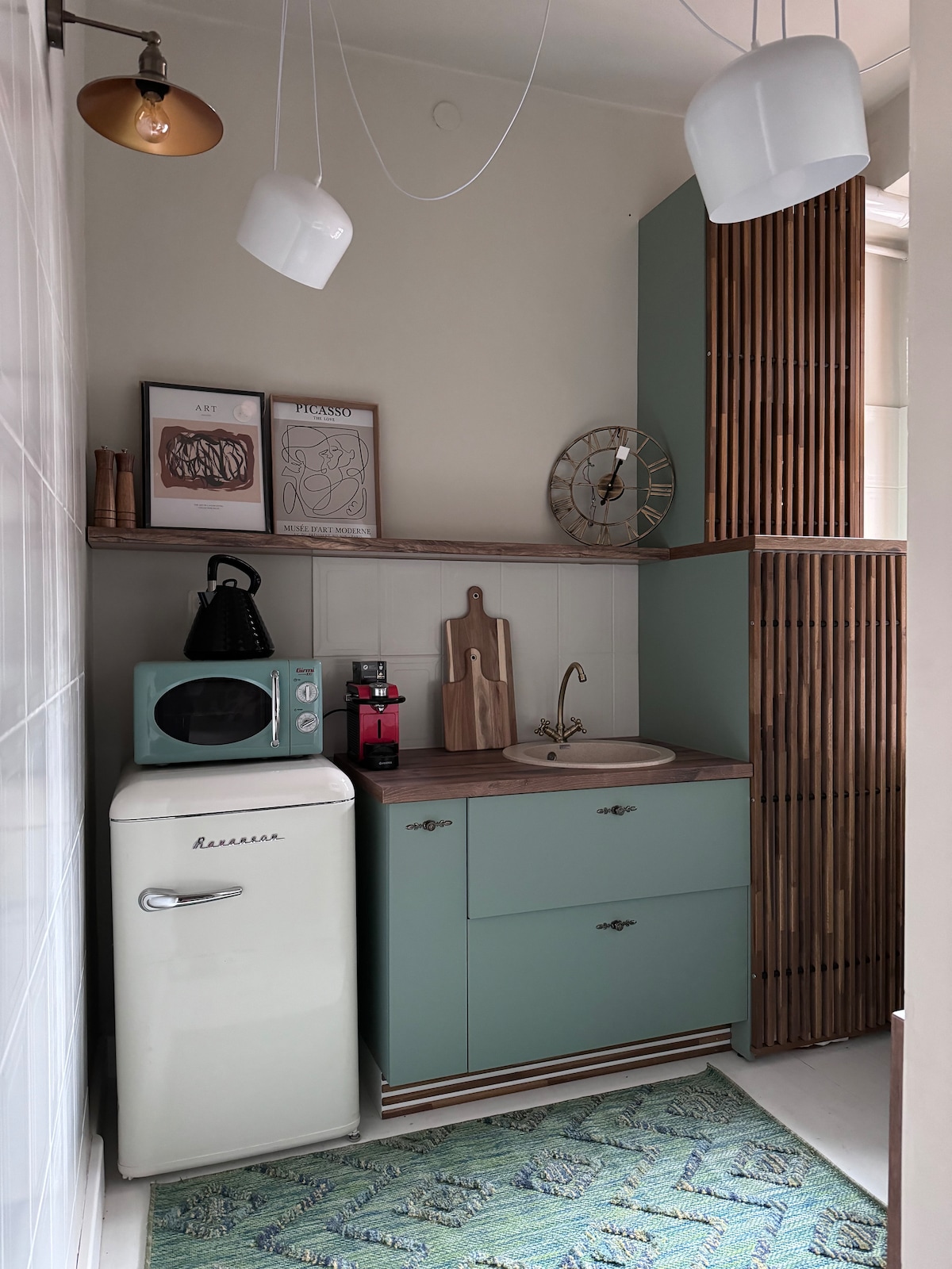 A compact kitchen features a vintage-style refrigerator in white. Soft green cabinetry complements a wooden countertop, while a sink is positioned under a decorative wall clock. A coffee maker and microwave are visible, with stylized art displayed on the walls and a colorful rug on the floor.