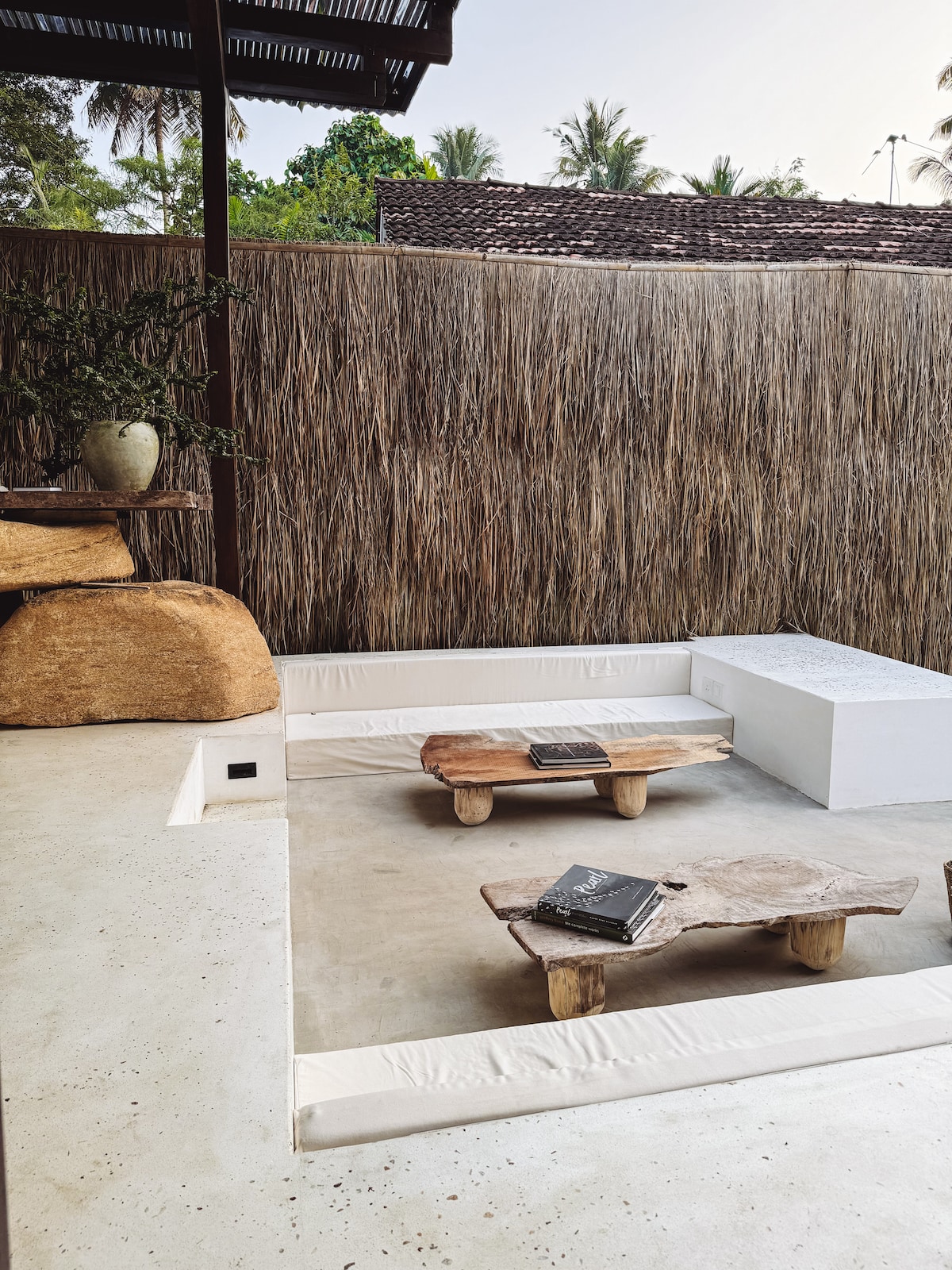 A serene outdoor seating area features natural stone and wood elements. A low wooden table sits between two cushioned benches, providing a relaxed atmosphere. Lush greenery is visible in the background, enclosed by a textured bamboo wall, enhancing the sense of privacy.