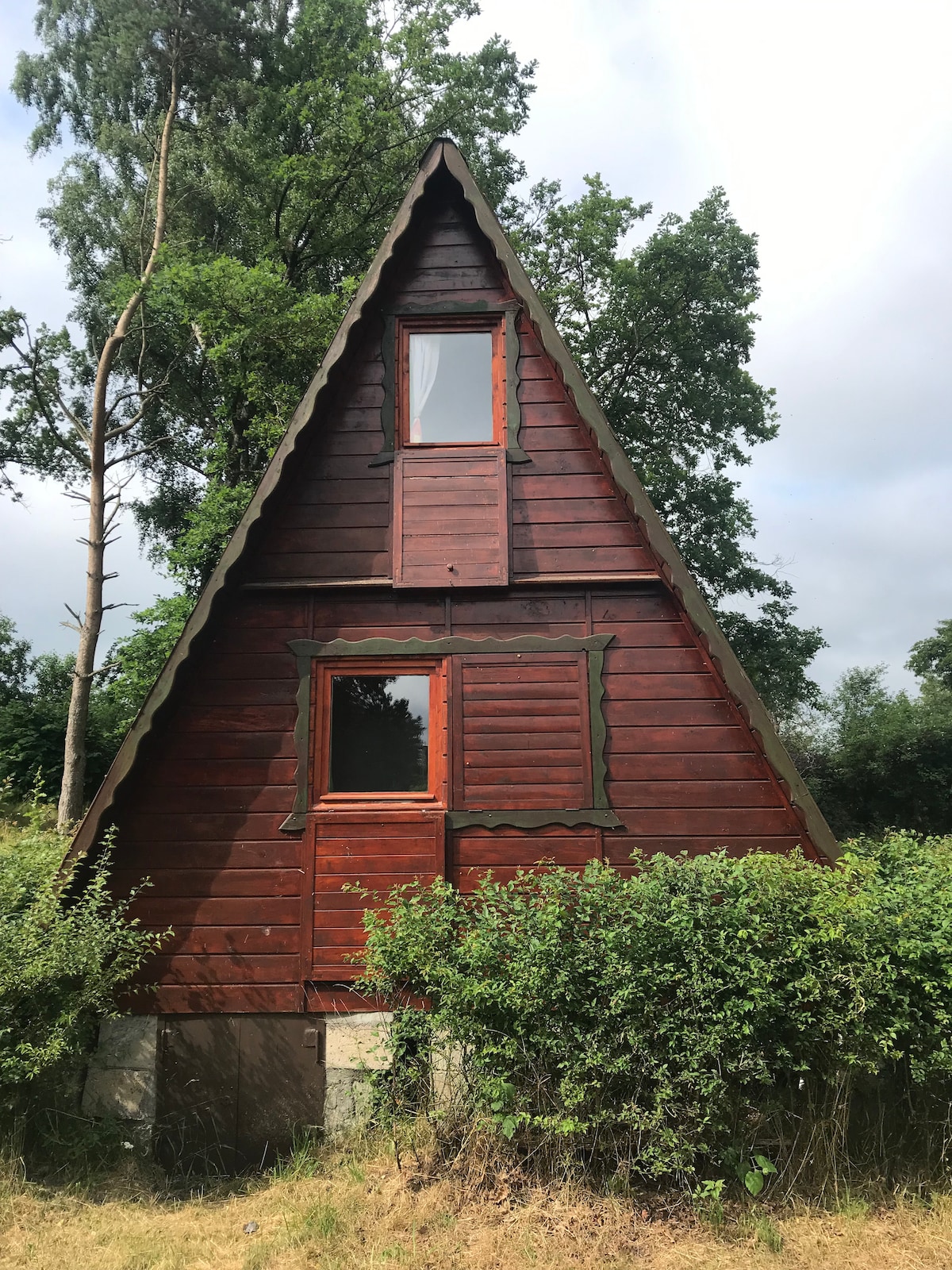A charming wooden A-frame cottage is framed by greenery. The structure features sloped roofs and multiple windows, providing a rustic appearance. A natural landscape surrounds the cottage, enhancing the inviting and serene setting.