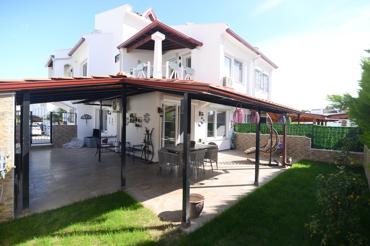 The outdoor area features a covered patio with a dining table and seating, surrounded by landscaped grass and stone walls. The villa showcases a welcoming design with multiple windows and balconies overlooking the garden, providing a bright and airy atmosphere.