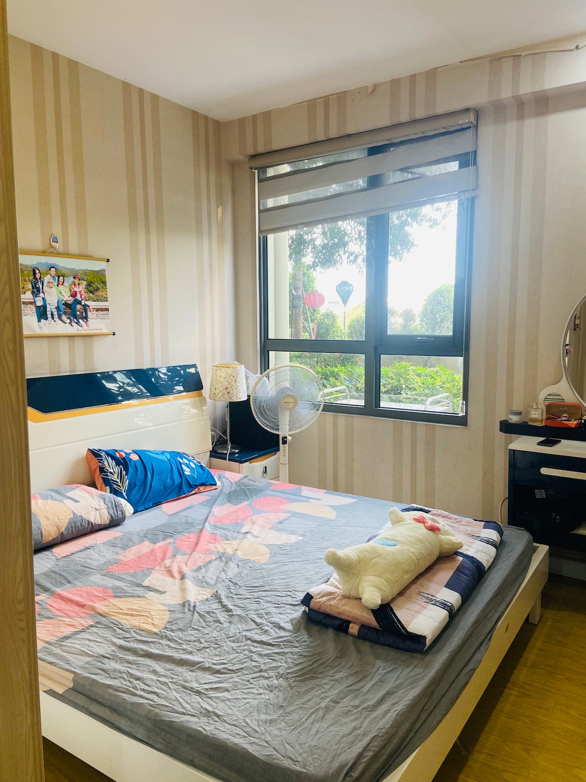 A cozy bedroom is illuminated by natural light from the large window, which overlooks greenery. The bed features a soft blanket with decorative pillows. A small fan and a vanity unit are present, adding functional elements to the space.