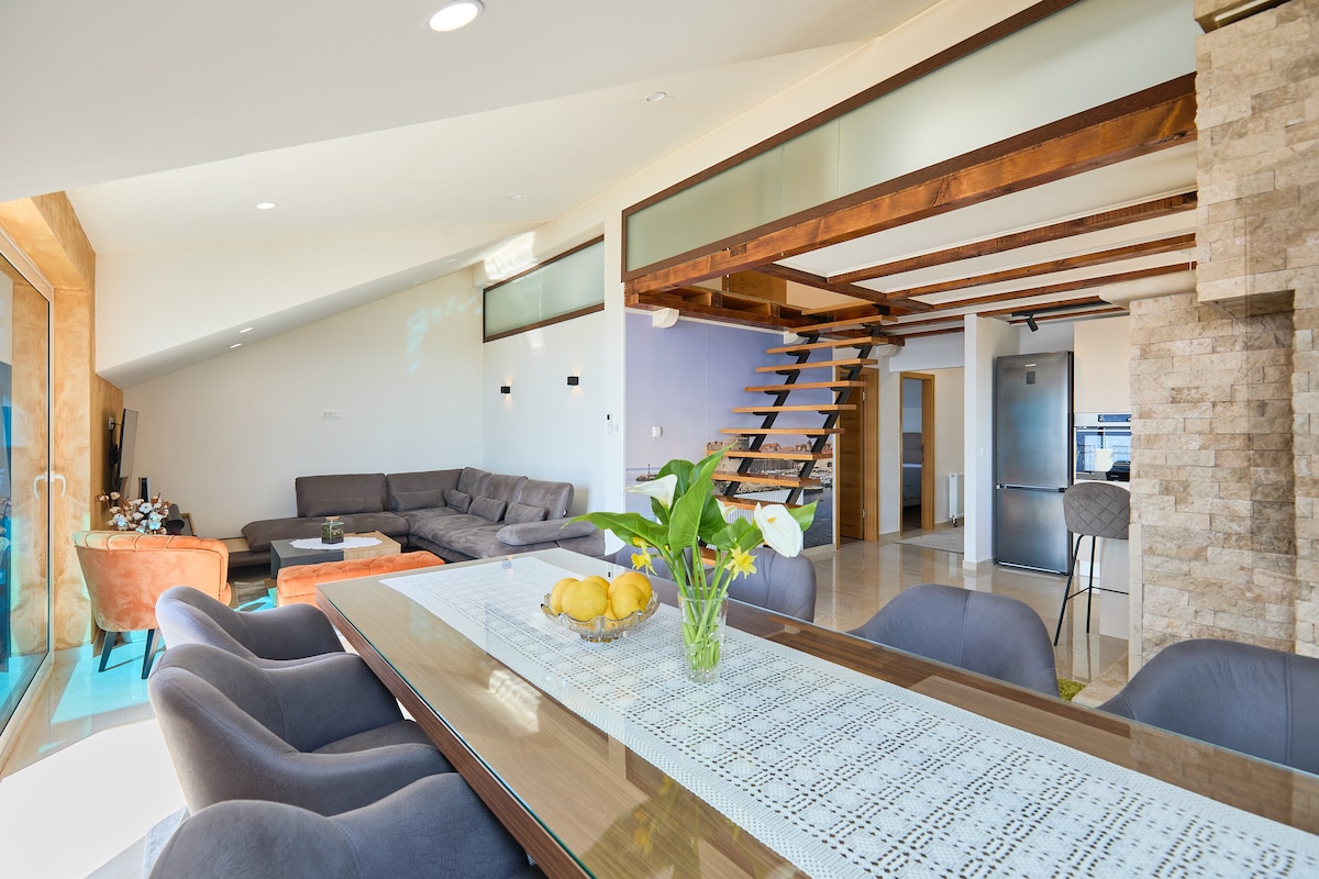 An open-plan living space features a modern dining table centered among plush, upholstered chairs. A sleek kitchen is visible alongside a comfortable sectional sofa. Natural light streams through expansive windows, illuminating the warm wood accents and textured stone details throughout the area.