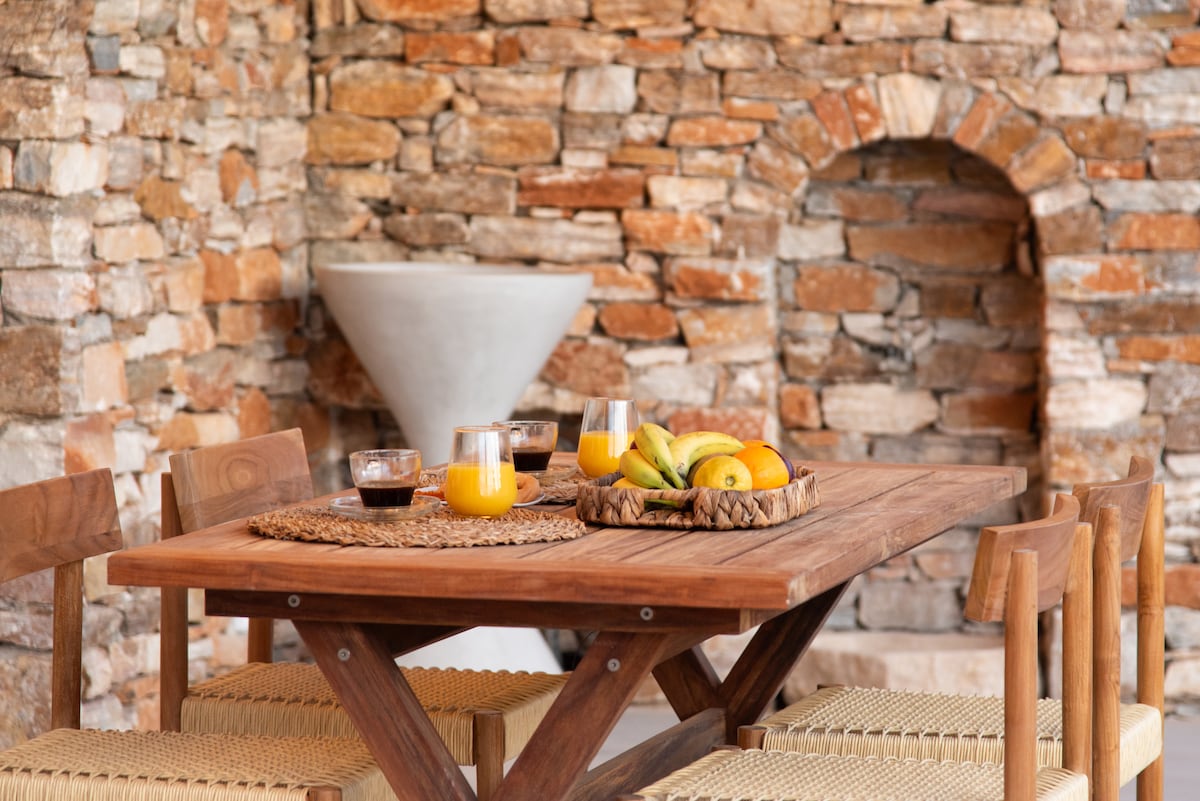 A wooden dining table is set with glasses of juice and coffee. A woven basket filled with fresh fruit sits in the center. The backdrop features rustic stone walls, adding a natural element to the dining area.