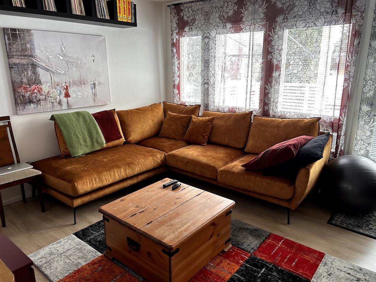 A spacious seating area is highlighted by a large, warm-toned sectional sofa adorned with colorful cushions. A rustic wooden coffee table sits centrally, with a remote control placed atop. Natural light filters through sheer curtains, creating a cozy and inviting atmosphere.