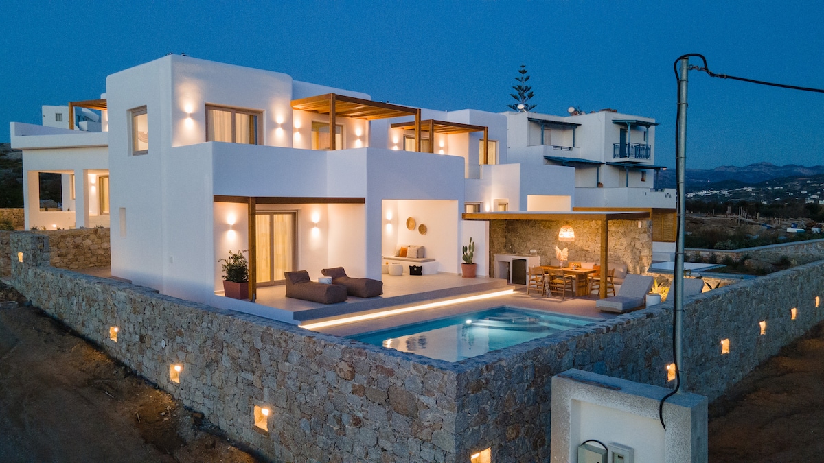 A modern two-story villa is captured at dusk, showcasing clean lines and a minimalist design. The exterior features large glass windows and a private pool illuminated by soft lighting, while a cozy seating area and dining space are visible under a covered patio.