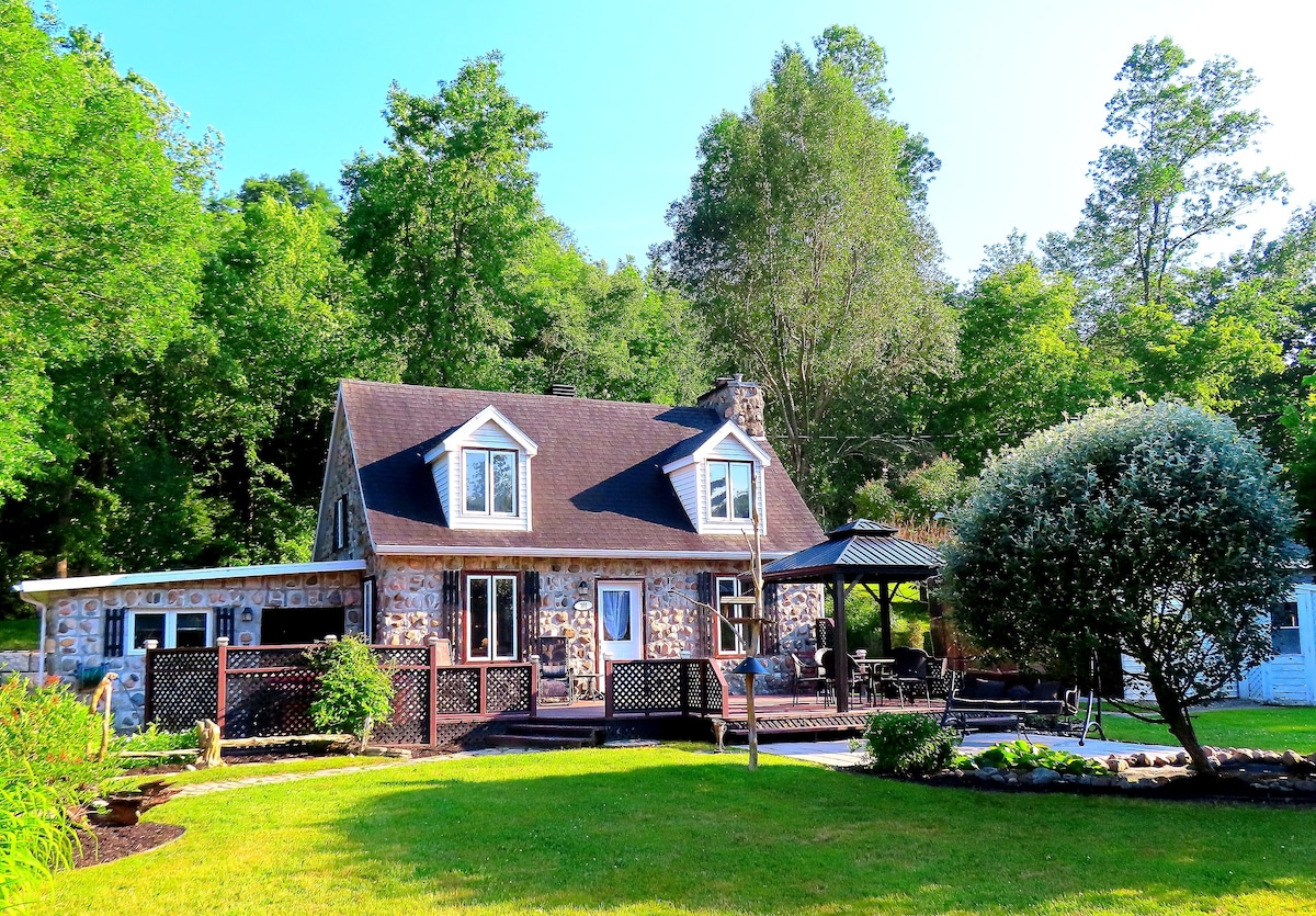 The exterior of a charming stone cottage is surrounded by lush greenery. A spacious deck offers seating and shade. Garden areas feature well-maintained landscaping, with a shrub and flower beds enhancing the outdoor setting. The structure reflects warm, inviting tones against the natural backdrop.