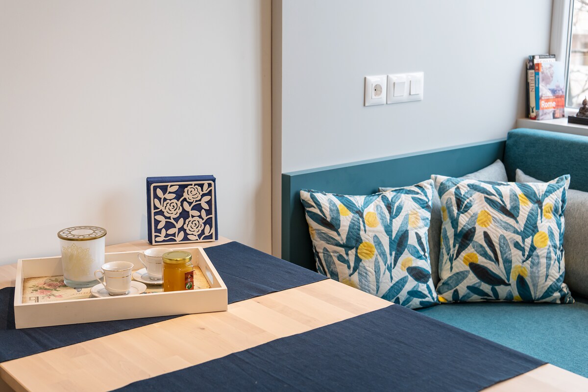 A dining table is set with a tray holding a cup, a small bowl, and a jar. A corner seating area features cushions adorned with floral patterns, complemented by a blue-green sofa. Natural light illuminates the cozy space.