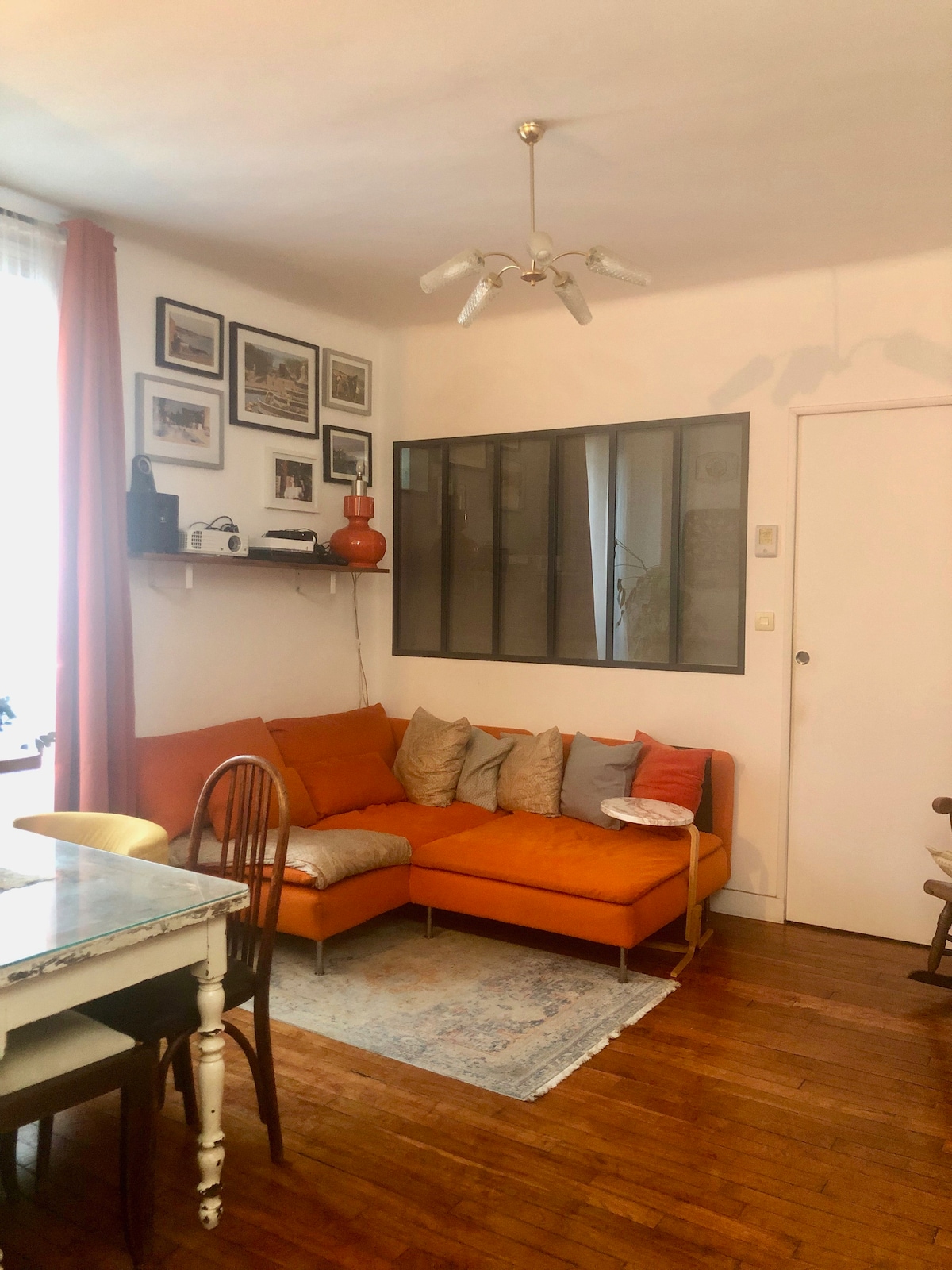 A cozy living area is featured with a bright orange sectional sofa adorned with multiple cushions. A minimalist glass partition separates the space, and framed photographs decorate the walls. A wooden dining table and chairs occupy one side, complemented by a softly patterned area rug.