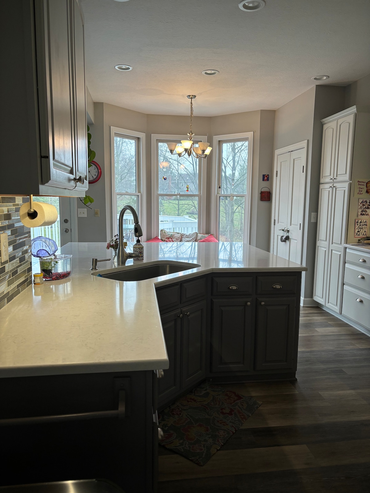 A modern kitchen is showcased with dark cabinetry and a contrasting light countertop. Natural light floods in from large bay windows, illuminating the space. A sink and faucet are situated on an island, with additional cabinetry and a pantry door visible.