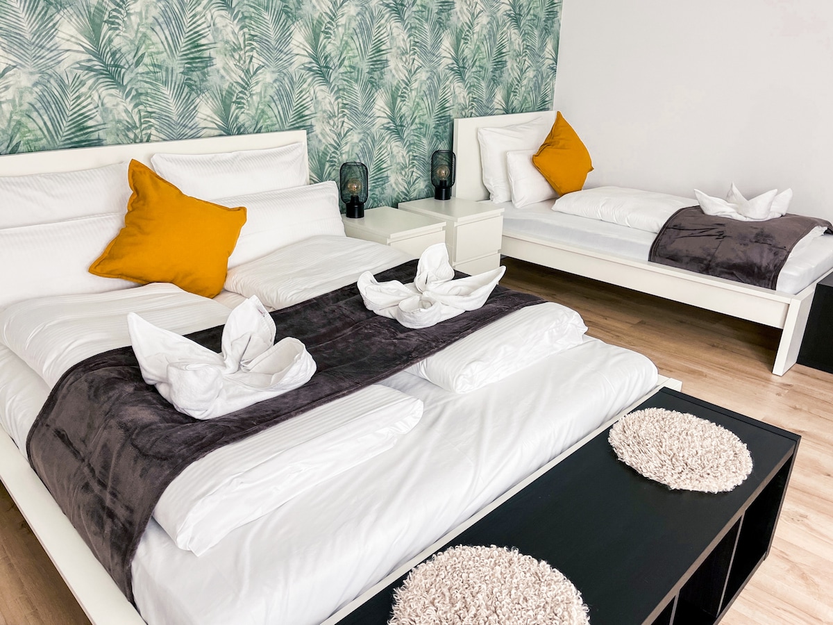 A sleeping area features two beds, one larger and one smaller, dressed in crisp linens. The wall behind is adorned with a leafy green wallpaper, complemented by two orange pillows on the larger bed. A bedside table sits between the beds, with decorative lamps providing soft lighting.