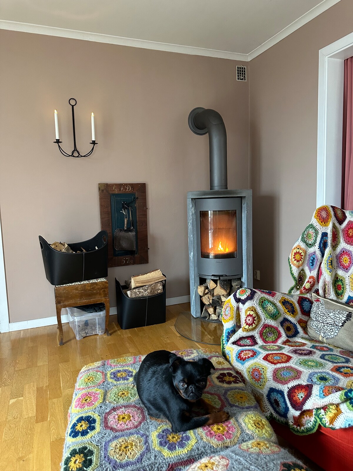 A cozy living area features a stone fireplace with a wood storage unit beside it. A patterned blanket covers a comfortable chair, and a dog rests nearby. Soft lighting is provided by wall sconces, and natural light fills the space through a window.