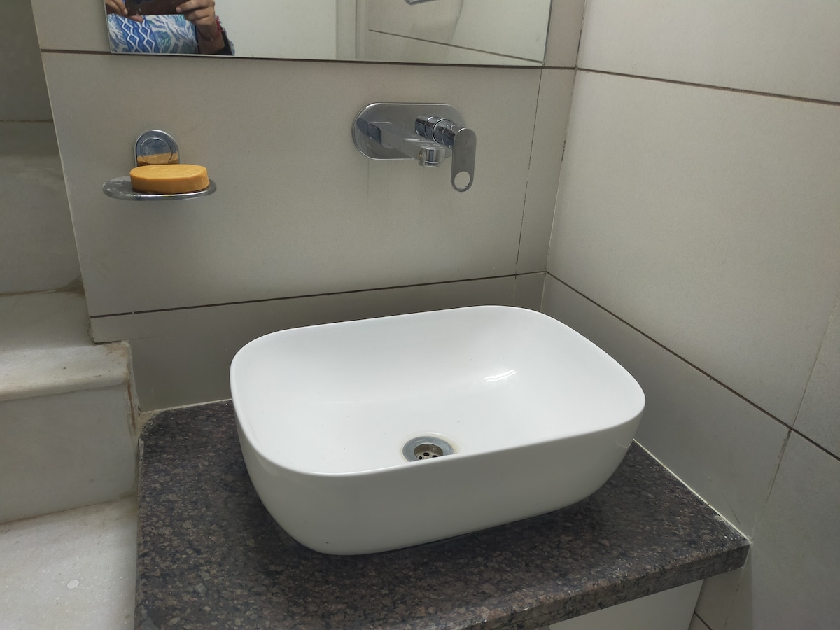 A modern bathroom sink is displayed on a dark granite countertop. The sink features a sleek, rounded design with a central drain. A wall-mounted faucet is positioned above it, and a small soap dish is mounted on the wall, holding a bar of soap.