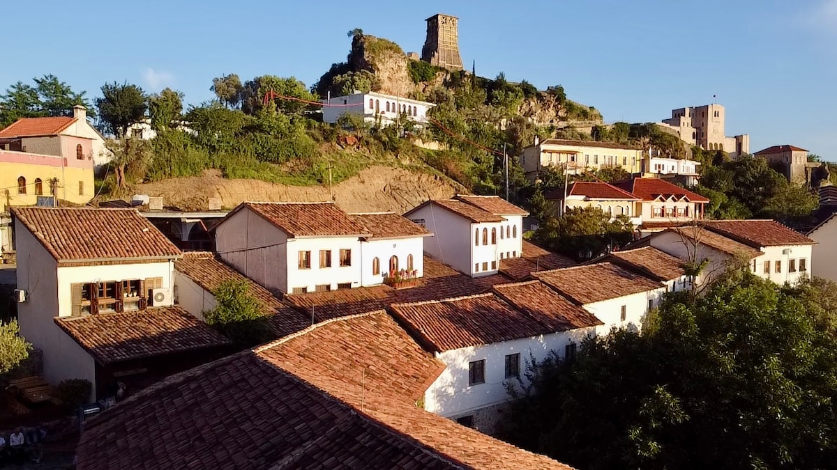 A panoramic view reveals traditional architecture featuring red-tiled roofs and whitewashed walls, nestled against a lush hillside. In the background, a historic castle stands prominently atop a hill, surrounded by greenery and older buildings, highlighting the charm of the Kruja Old Bazaar area.
