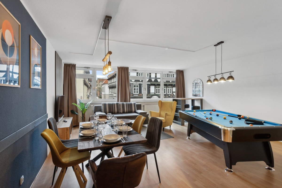 A combined living and dining area features a large dining table set with plates and glasses, complemented by stylish yellow and dark chairs. A sleek sofa invites relaxation, while a blue pool table adds a playful element. Large windows provide natural light, showcasing views of nearby architecture.
