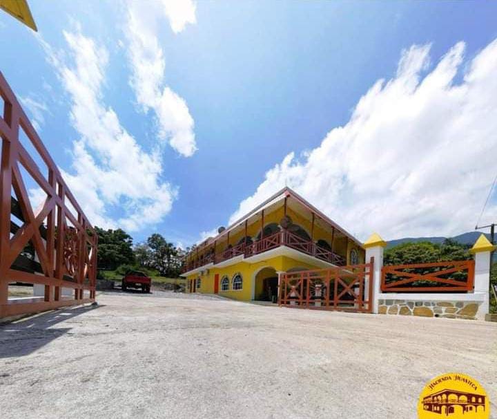 A welcoming exterior view of a vibrant yellow building is captured, set against a blue sky with scattered clouds. The entrance is lined with a wooden fence, and a red car is parked nearby, adding to the inviting atmosphere of the property.