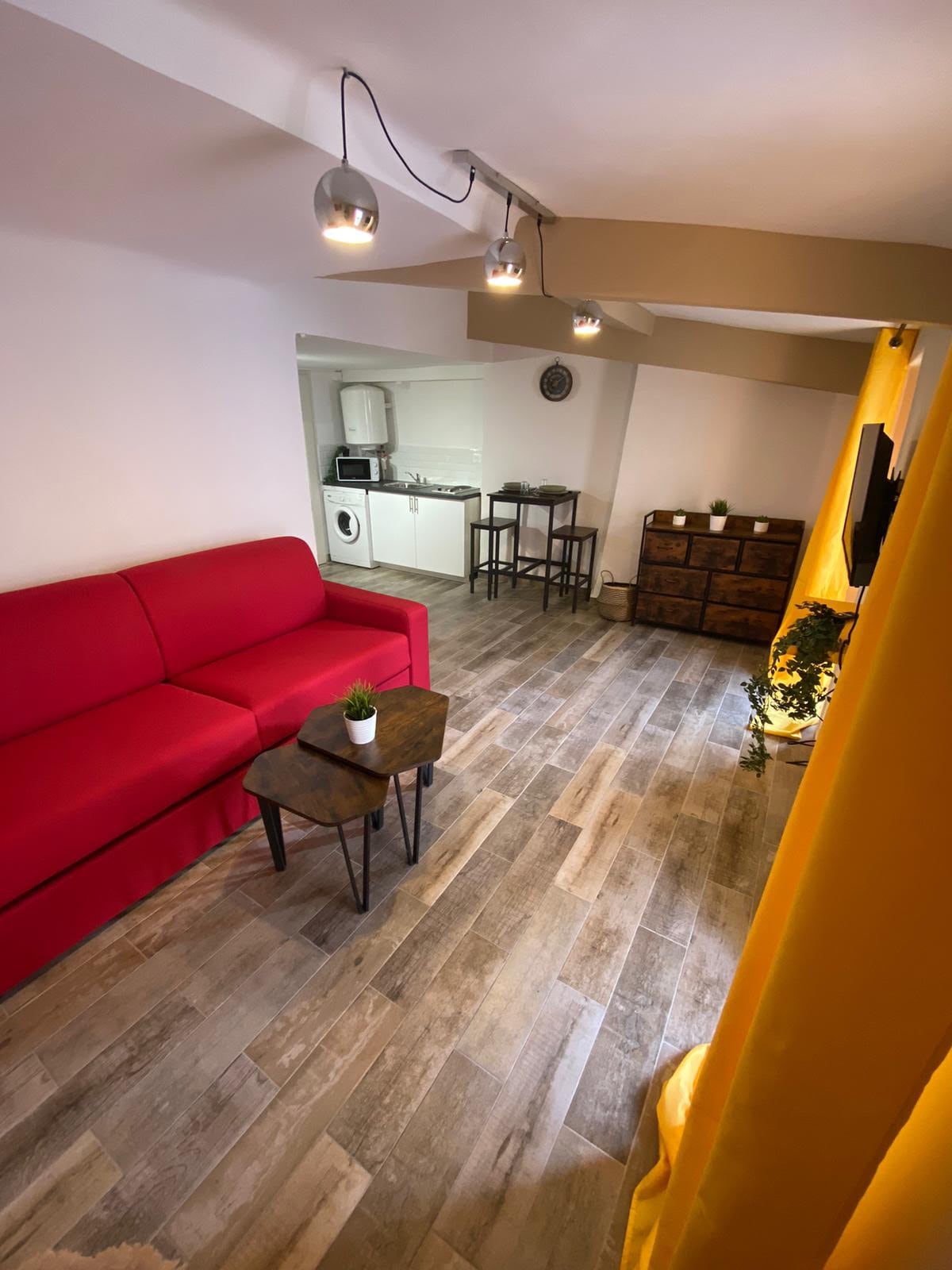 A bright living area is furnished with a red sofa, accompanied by two small tables. Warm wooden flooring complements the space, which is illuminated by track lighting. The area is partially enclosed by yellow curtains, with a glimpse of a kitchen and dining area in the background.