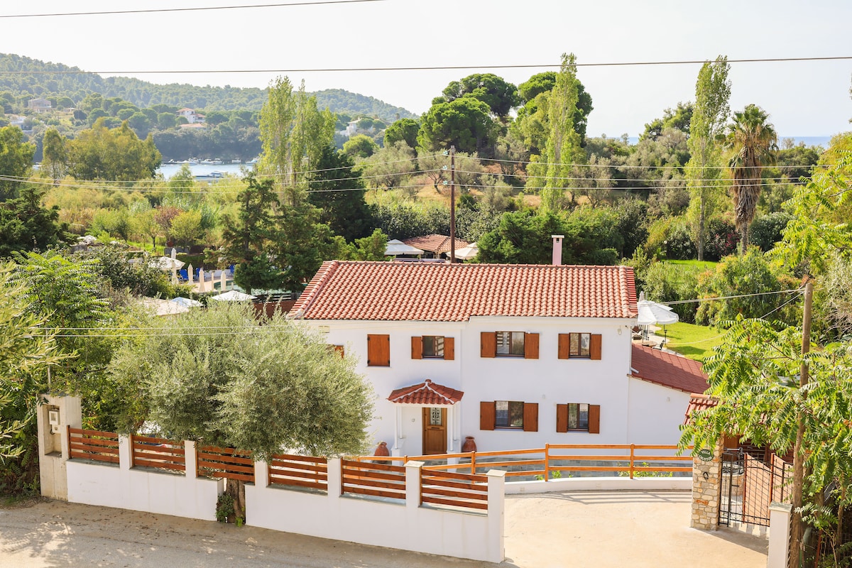 A two-story villa is positioned among lush greenery, featuring a red-tiled roof and white exterior. The surrounding landscape includes trees and distant views of water. A fenced area with wooden accents provides a sense of privacy, while the outdoor space seamlessly integrates with nature.