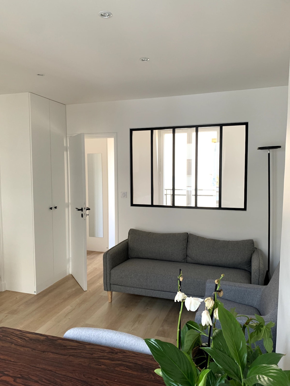 A simple living area features a gray sofa positioned against a wall, with light wood flooring. A small plant with white flowers is visible in the foreground. A doorway is present, leading to another room, framed by a large window-like partition that adds brightness.