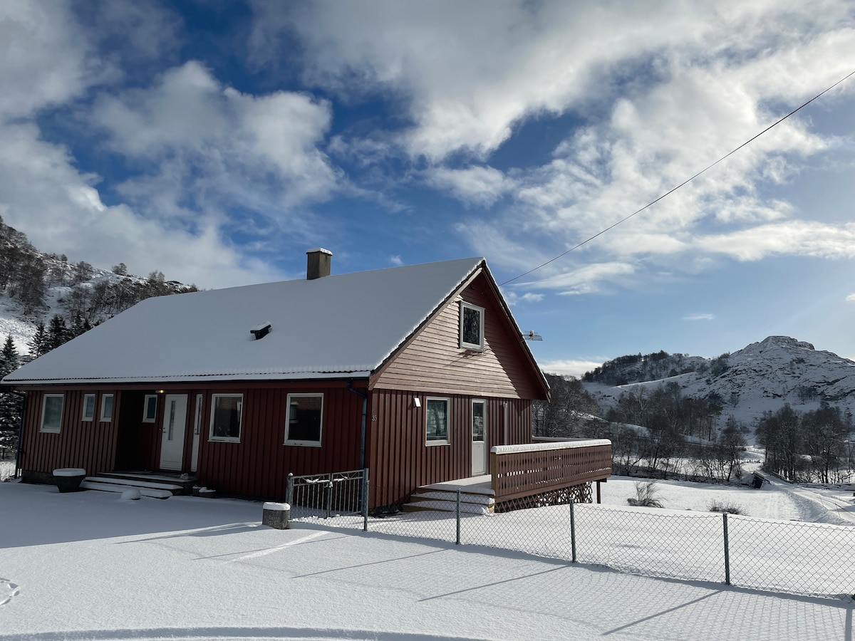 A red wooden house is set against a backdrop of snow-covered terrain and rolling hills. The structure features a front deck with steps leading to a fenced area. A clear blue sky with scattered clouds enhances the tranquil winter scene.