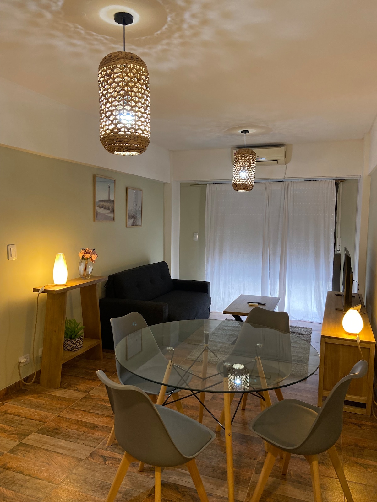 A dining area is illuminated by woven pendant lights, featuring a round glass table surrounded by four light grey chairs. A black sofa is positioned against the wall, and a television cabinet provides a subtle contrast. Natural light filters through sheer white curtains.