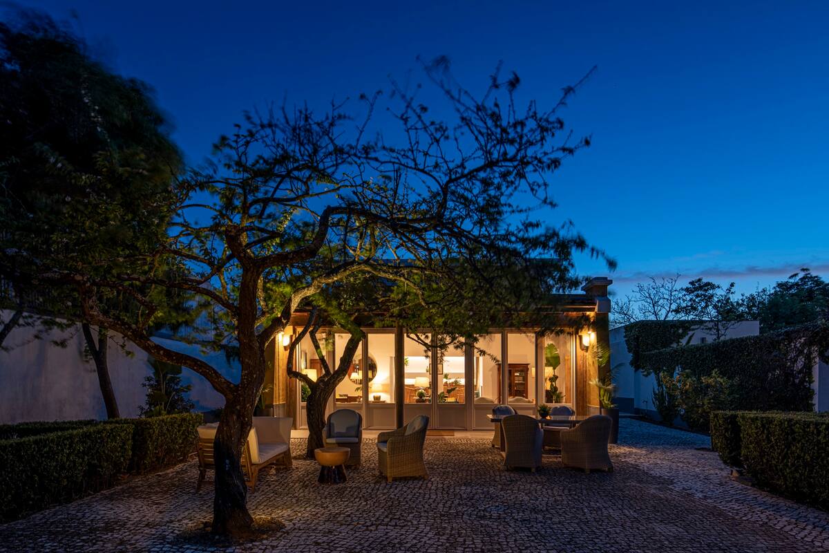 An outdoor area is illuminated by soft lighting at dusk, showcasing a cozy seating arrangement with wicker furniture surrounded by well-manicured shrubbery. A tree stands prominently, adding texture to the serene space, while large glass doors reveal the interior of the villa.