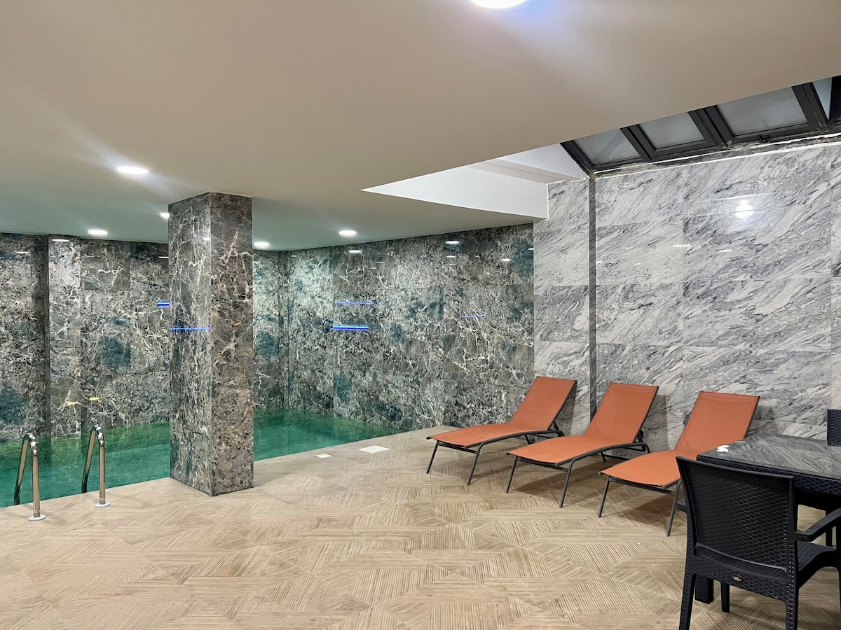 An indoor pool area is surrounded by elegant marble walls, creating a serene atmosphere. Lounge chairs in warm orange are positioned nearby, inviting relaxation. Subtle lighting enhances the tranquil environment, while the pool water reflects the surrounding marble textures.