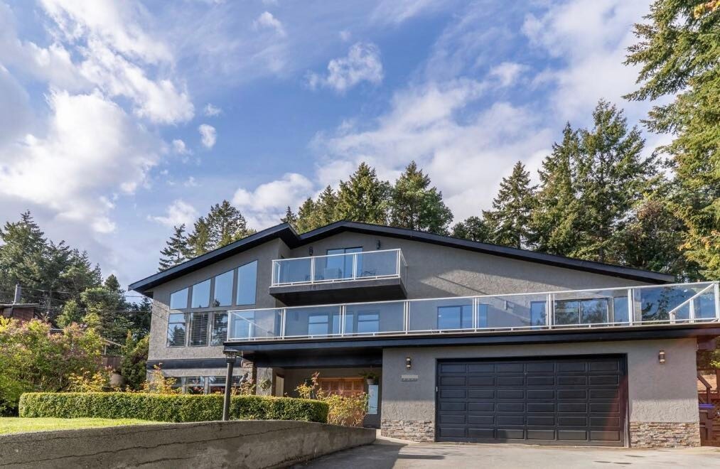 The exterior of the home features a modern design with large glass balcony railings and a double garage. The structure is surrounded by lush trees and landscaping, set against a bright sky with scattered clouds.