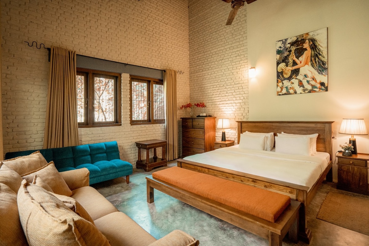 An open concept room features a kingsized bed and sofa bed, complemented by wooden furnishings. Two windows allow natural light and provide views of the outdoors. A textured wall and soft lighting create a calming environment, while a vibrant wall art piece adds a touch of color.