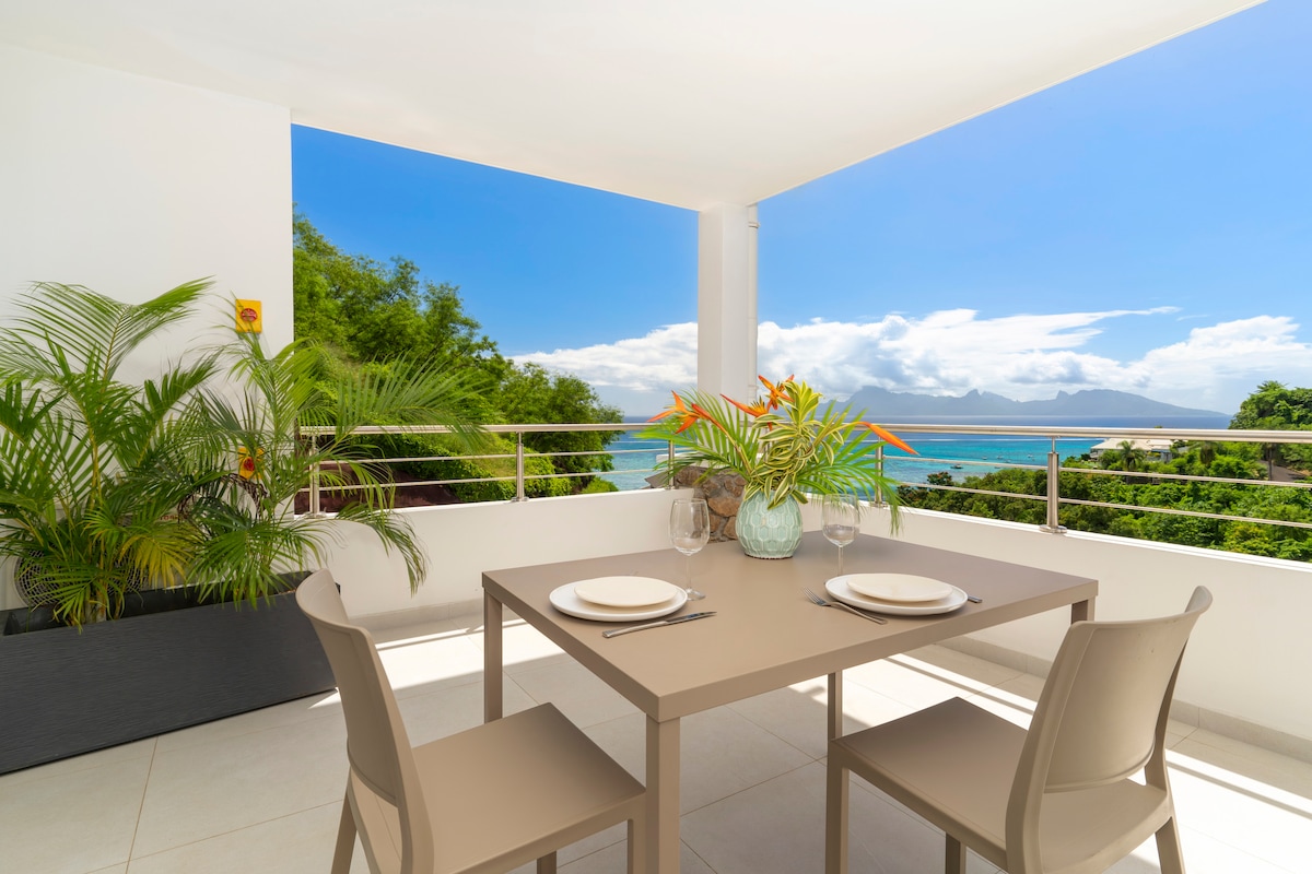 A terrace is shown with a dining table set for two, accompanied by stylish chairs. Lush greenery is visible in a planter, enhancing the tranquil atmosphere. The ocean and distant mountains are framed by the open balcony, inviting relaxation and enjoyment of the scenic views.