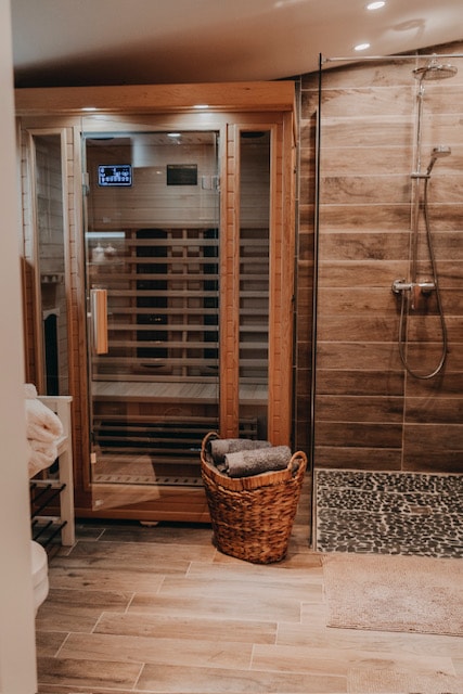 A modern bathroom features a wooden infrared sauna enclosed in glass, alongside a rainfall shower. Textured stone tiles line the shower area, and a woven basket holds bath towels. Soft lighting accents the warm wooden tones and neutral surfaces.
