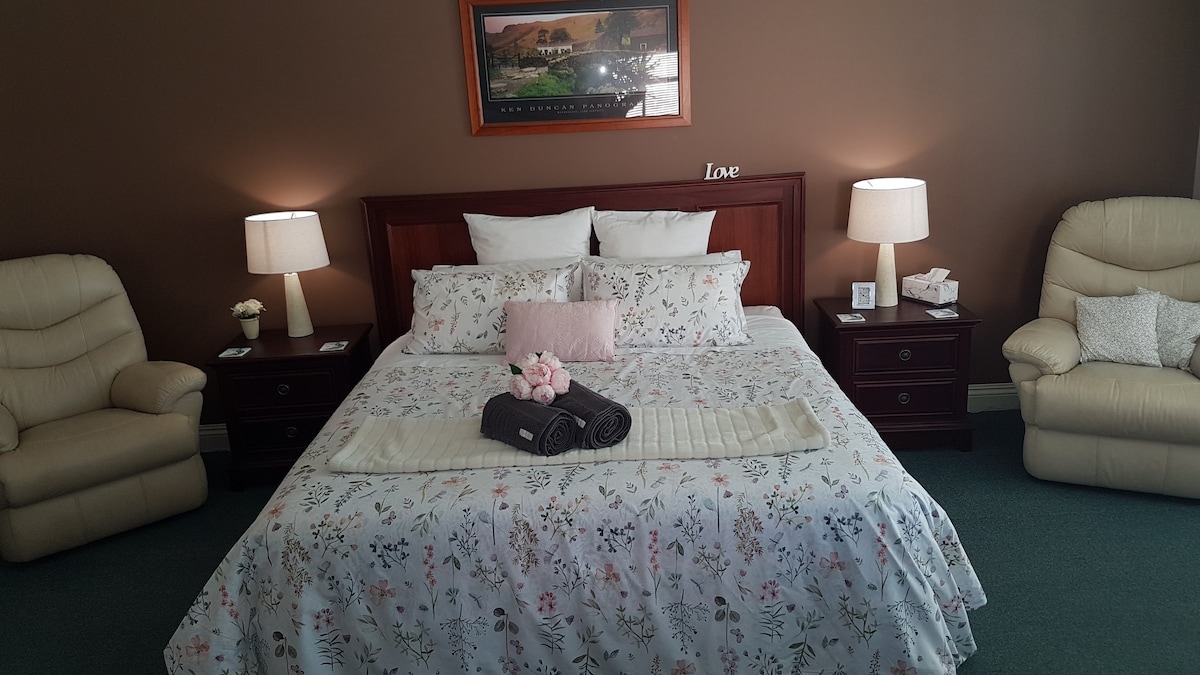 A spacious bedroom is highlighted by a king-size bed adorned with a floral-patterned blanket. Two bedside tables with lamps flank the bed, while two light-colored armchairs provide additional seating. Soft light illuminates the room, complemented by neutral-toned walls and a framed landscape print.