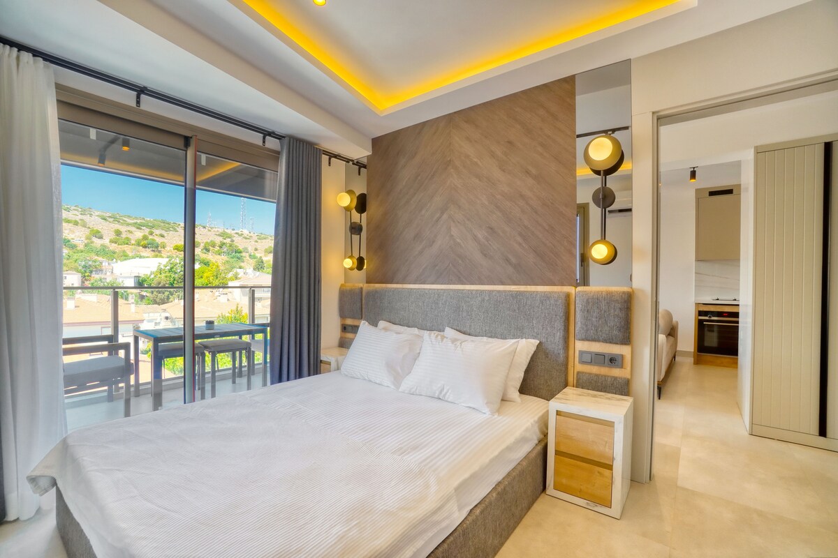 A spacious bedroom features a modern design, with a wide bed dressed in crisp linens. Natural light floods in through large glass doors, leading to a balcony with outdoor seating. Soft lighting fixtures enhance the contemporary aesthetic of the room.