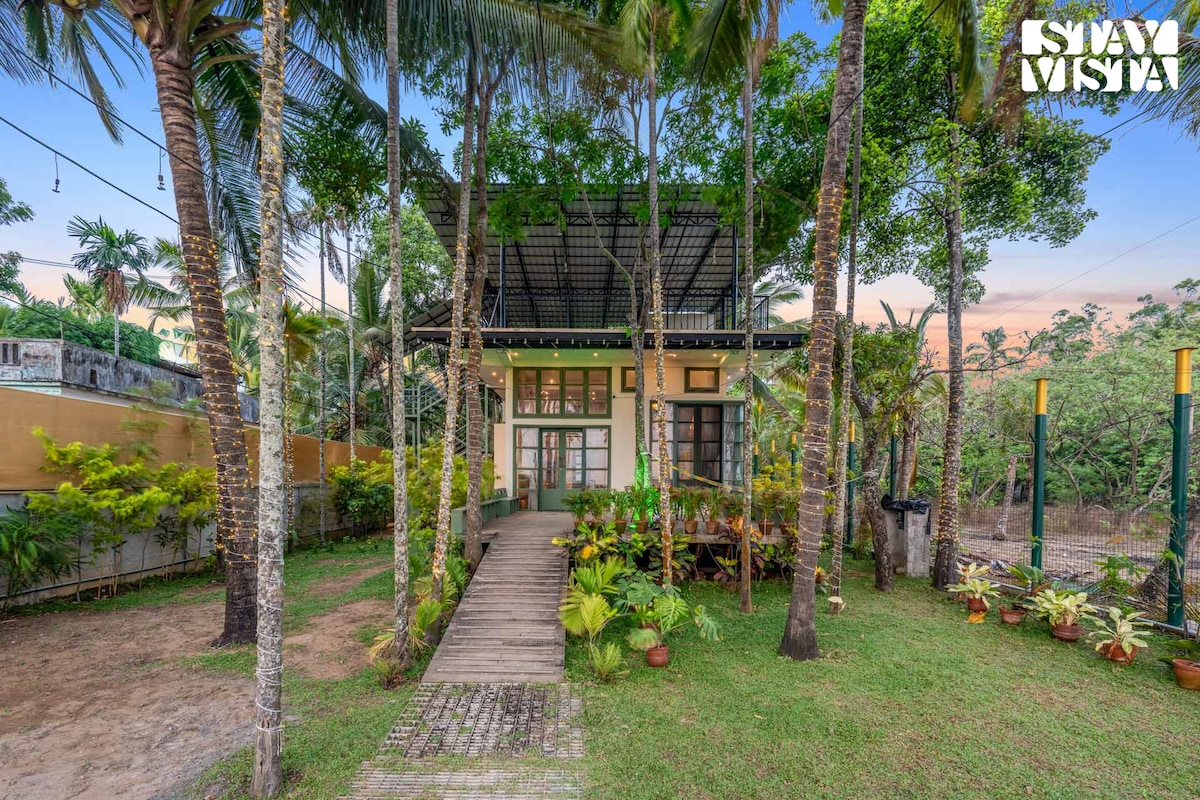A charming two-story villa is surrounded by lush greenery and palm trees. A wooden pathway leads to the entrance, which features large windows that enhance natural light. The outdoor space is filled with well-maintained plants, creating a tranquil and inviting environment.