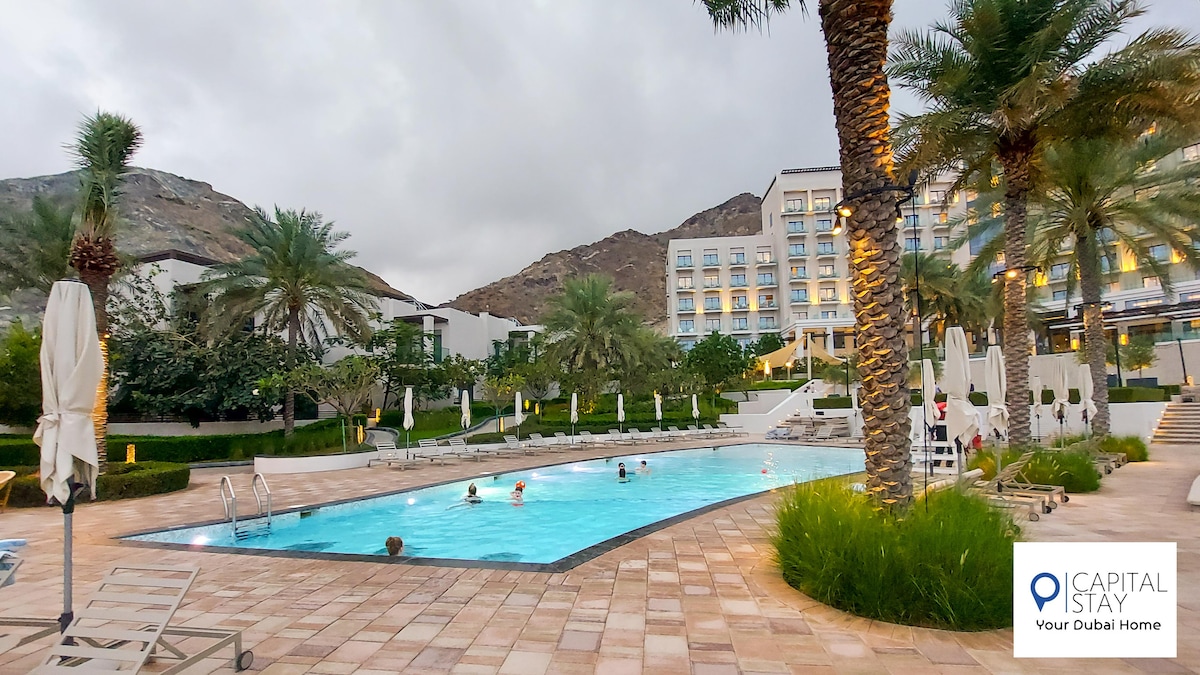 An outdoor pool area is surrounded by palm trees and lounge chairs. Guests can be seen enjoying the pool, with mountains in the background. The sky is overcast, adding a serene ambiance to the setting.