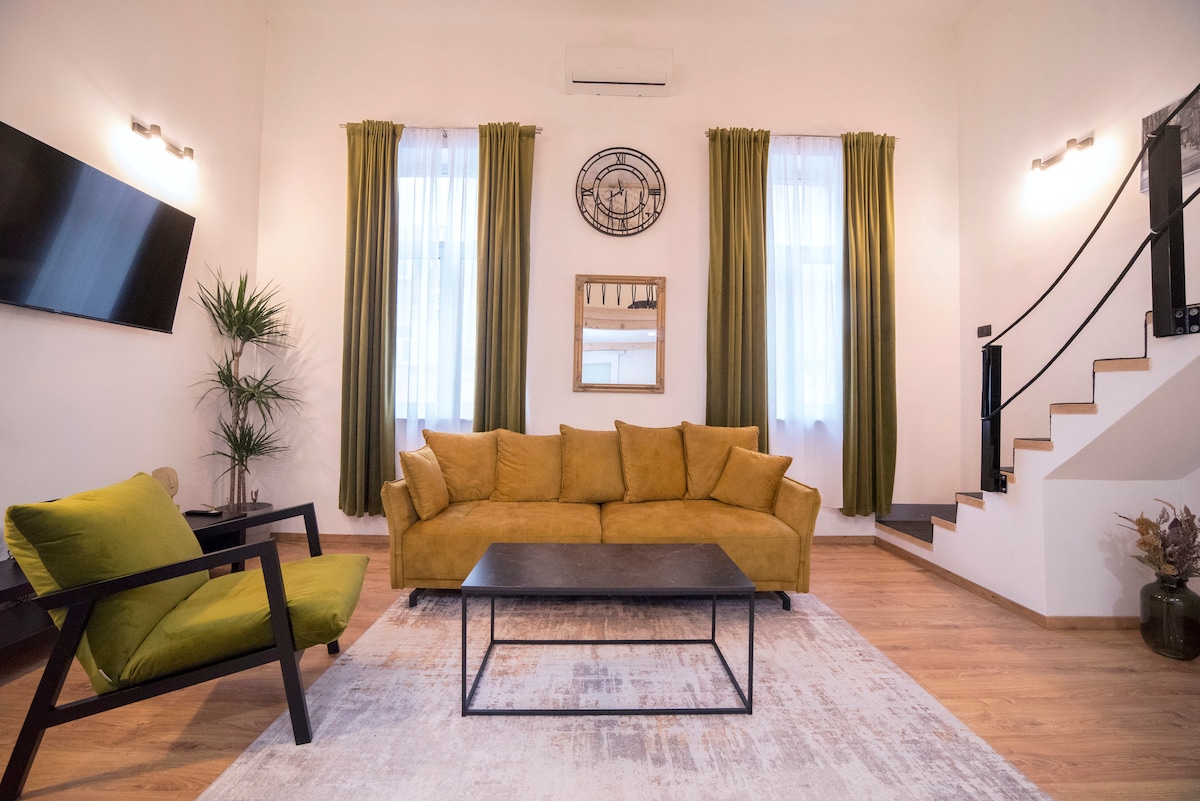 A spacious living area is presented with a plush yellow sofa and a black coffee table. Two tall windows provide natural light, complemented by long, green curtains. A modern television is mounted on the wall, and a decorative mirror adds depth to the inviting space.