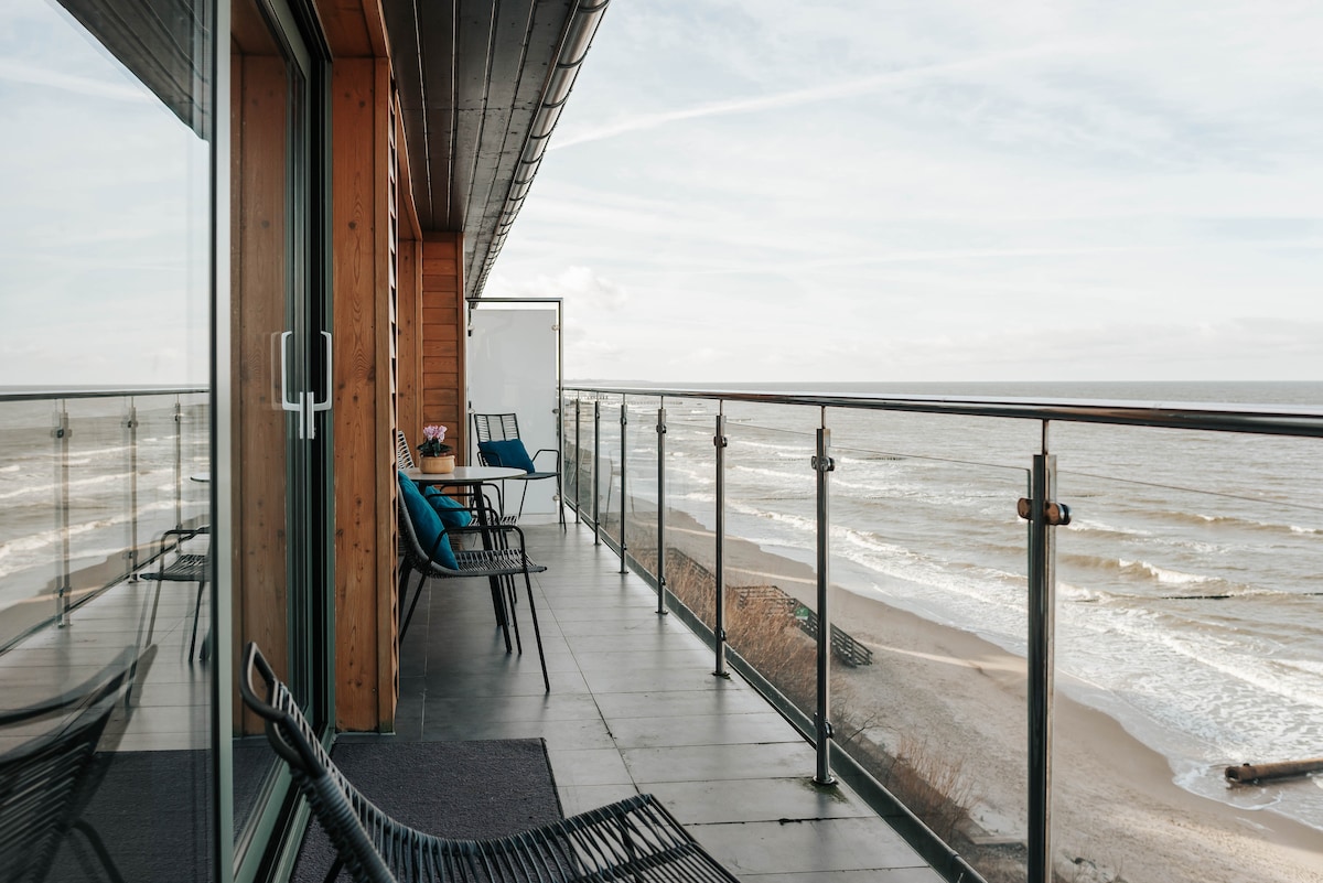 A spacious balcony offers uninterrupted views of the beach and ocean, with glass railings enhancing the open feel. Metal chairs and a small table provide seating options, while the wooden siding of the building complements the coastal scenery.