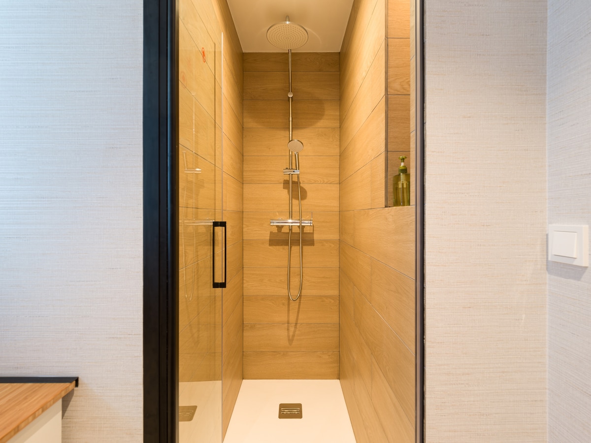 A spacious, walk-in shower is outlined by sleek glass doors. The walls are covered in warm wood-like tiles, creating a natural ambiance. A modern showerhead is mounted above, and shelves are visible for toiletries, enhancing the practical design.