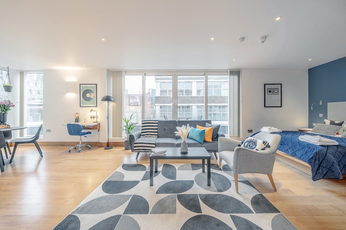 The spacious living area is furnished with a grey sofa and two chairs, while a modern desk and lamp are positioned near a large window. A textured rug underlines the seating arrangement, and a king-size bed is visible in the corner, accented by decorative pillows.