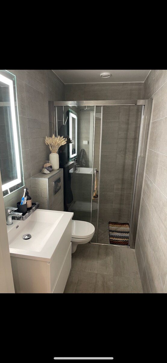 A modern bathroom is presented, showcasing a sleek white vanity with a basin and decorative items. A glass shower enclosure is visible, with neutral-toned tiles adorning the walls and floor. Soft lighting from the mirror illuminates the space, enhancing the clean and organized appearance.