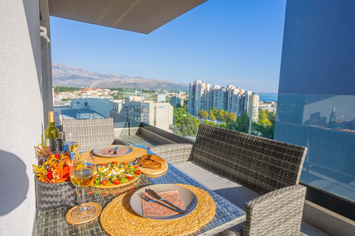 A balcony is furnished with a table set for dining, featuring plates of food, glasses, and a bottle. Two woven chairs provide comfortable seating. The view includes distant mountains and buildings under a clear blue sky, with a glimpse of the sea in the background.
