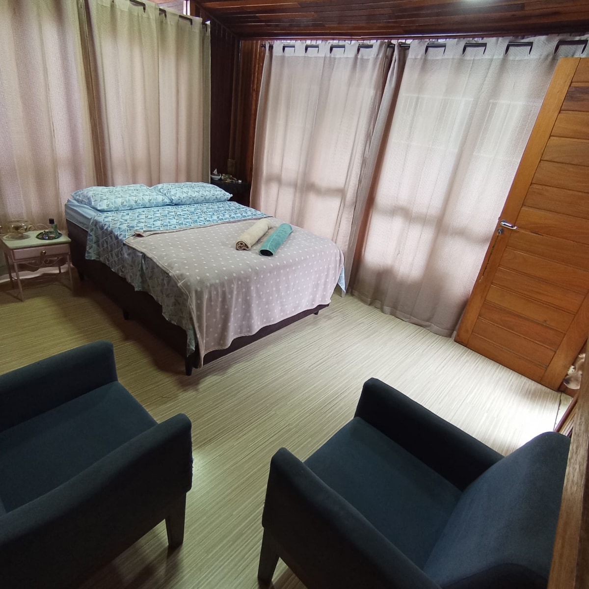A spacious sleeping area features a light-colored polka dot bedspread covering a large bed, with blue patterned pillows positioned neatly. Two dark armchairs are placed beside the bed, and natural light filters through sheer curtains, creating a soft ambiance.