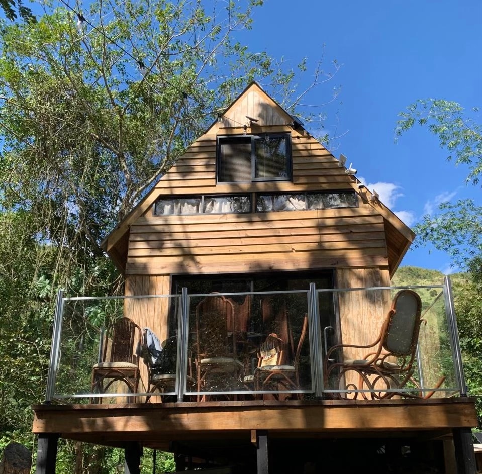 The cabin features a wooden exterior with large windows allowing natural light. A spacious deck is furnished with several chairs, offering a relaxing space surrounded by greenery. The clear sky enhances the tranquil atmosphere of the riverside setting.